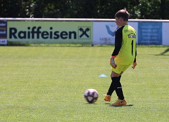 Ein junger Fußballspieler in einem gelben Trikot tritt einen Fußball auf einem Feld, auf dem ein Raiffeisen-Banner im Hintergrund steht.