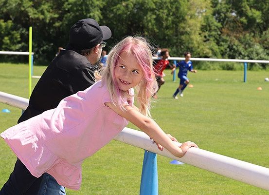 Ein junges Mädchen mit rosa Haaren lehnt sich an einen weißen Pfosten auf einem Rasenfeld. Ein Mann in einem schwarzen Hut steht in der Nähe. Im Hintergrund spielen zwei Personen Fußball.