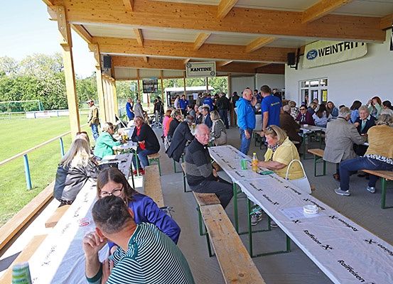 Eine Gruppe von Menschen sitzt auf Bänken in einem überdachten Bereich mit Holzbalken und genießt Essen und Getränke. Einige tragen Brillen, und eine Frau hat eine Handtasche. Ein grünes Feld ist draußen sichtbar.