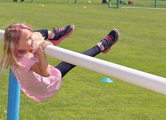 Ein kleines Mädchen hängt an einer horizontalen Stange und lächelt, wahrscheinlich auf einem Spielplatz.