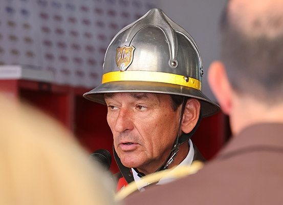 Ein Feuerwehrmann mit Helm spricht in ein Mikrofon, wobei ein Mann in Uniform vor einem roten Hintergrund zuhört.