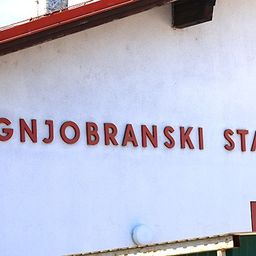 Ein weißes Gebäude mit rotem Dach hat ein Schild, das 'OGNJOBRASKI STAN' besagt. Unter dem Schild befindet sich eine Lampe und ein kleines Logo.