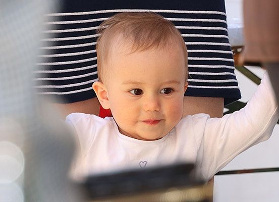 Ein Baby sitzt auf dem Schoß einer Person und trägt ein weißes T-Shirt mit Herzdesign, schaut nach vorne.