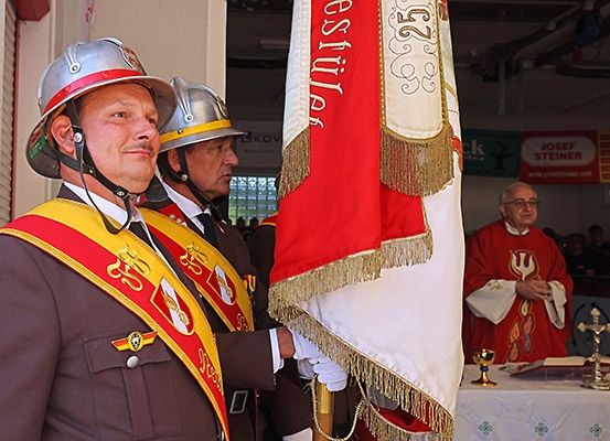 Zwei Feuerwehrleute in Uniform stehen neben einer Fahne in einer Kirche. Ein Priester steht an einem Tisch mit einem Buch und einem Kelch.