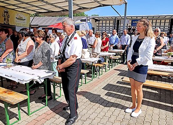 Eine Menschenmenge versammelt sich um Tische, einige tragen Sonnenbrillen. Ein Mann mit einer Kamera steht vor einer Frau in formeller Kleidung.