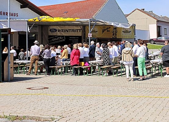 Viele Menschen stehen in einer Schlange vor einem Biergarten, in der Nähe von Reihen von Bänken und Tischen. Ein Zelt mit einem gelben Baldachin ist in der Nähe aufgebaut.