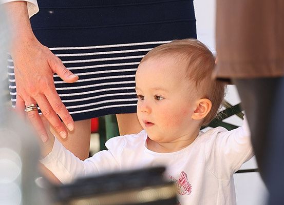 Ein kleines Baby in einem weißen T-Shirt streckt sich in einem unscharfen Hintergrund in Richtung einer Hand mit einem Ring aus.