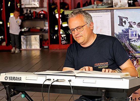 Ein Mann sitzt an einem Yamaha-Keyboard mit einem offenen Buch vor ihm. Er trägt eine Brille und ein schwarzes T-Shirt. Hinter ihm steht ein Kind in der Nähe einer Feuerwehruniform.