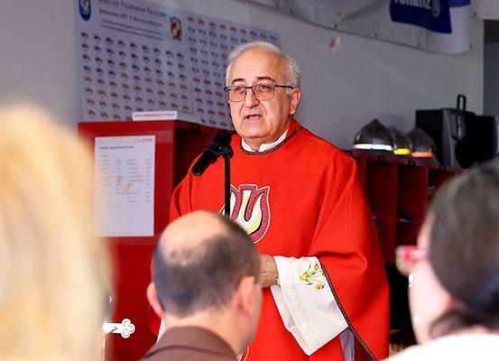 Ein älterer Priester in einer roten Robe spricht in ein Mikrofon, umgeben von mehreren zuhörenden Personen. Hinter ihm befindet sich eine Tafel mit Text und Zahlen.