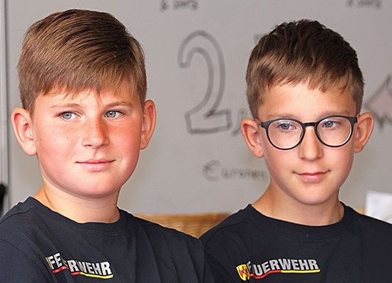 Zwei Jungen in identischen schwarzen T-Shirts mit dem Aufdruck 'Feuerwehr', die für ein Foto posieren. Ein Junge trägt eine Brille. Hinter ihnen befindet sich eine Tafel mit der Nummer '2' und einem Logo.