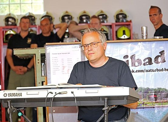 Ein Mann spielt ein Yamaha-Keyboard und schaut in eine Notensammlung, während drei andere Männer in schwarzen Hemden und mit Uhren im Hintergrund stehen.
