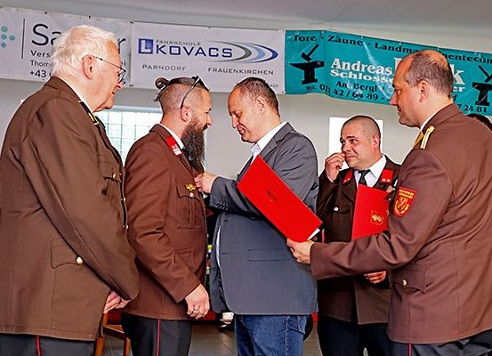 Fünf Männer in Uniformen, zwei mit roten Ordnern, einer erhält eine Medaille, und zwei andere beobachten. Hinter ihnen Banner mit Logos und Text.