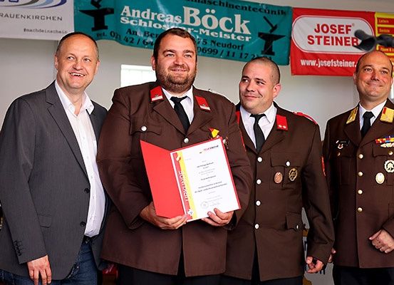 Vier Männer stehen zusammen, einer hält ein Zertifikat in der Hand und trägt braune Uniformen mit Medaillen. Sie stehen vor einem Banner mit Text, das Andreas Bock Schlossermeister liest.