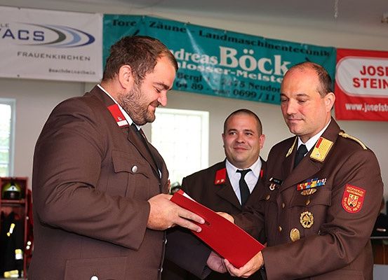 Drei Männer in Militäruniformen stehen drinnen. Einer hält eine rote Mappe, und die anderen beiden schauen darauf. Hinter ihnen befinden sich Banner und ein Regal mit Feuerschutzausrüstung.