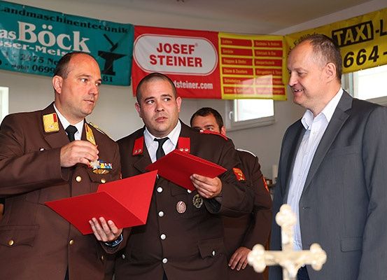 Drei Männer in Uniformen, einer präsentiert eine Auszeichnung, stehen vor einem Banner und einem Fenster. Ein weiterer Mann in einem Anzug steht in der Nähe und lächelt.