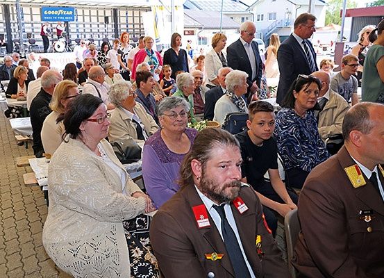 Eine Gruppe von Menschen, darunter ältere Erwachsene, sitzt in einer Reihe, wahrscheinlich bei einer Veranstaltung. Sie tragen alle formelle Kleidung. Vor ihnen steht eine Person, und links ist ein Tisch platziert.
