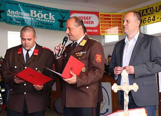 Drei Männer in Uniformen stehen vor einem Mikrofon und Dokumenten. Ein Mann liest aus einer roten Mappe, während ein anderer ein Mikrofon hält und spricht. Ein Mann in einem Anzug hält ein Kreuz vor ihnen. Hinter ihnen sind Banner mit Firmenlogos zu sehen.