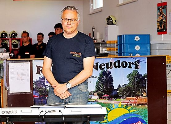 Ein Mann steht vor einem Yamaha-Keyboard, mit mehreren Leuten dahinter, die Helme tragen. Hinter dem Mann befindet sich ein Banner mit einem Strandbild.