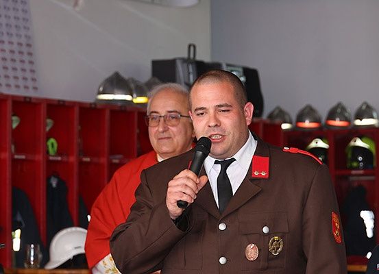 Ein Mann in einer braunen Militäruniform spricht in ein Mikrofon, während ein anderer Mann in einem roten Mantel hinter ihm steht. Der Raum ist mit roten Spinden und Helmen ausgestattet.
