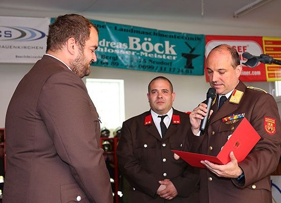 Drei Männer in Militäruniformen, einer hält ein Mikrofon und liest, andere stehen und schauen zu ihm, mit einem Banner im Hintergrund.