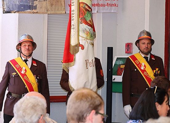 Zwei Männer in Uniformen mit Schärpen und Helmen stehen vor einer Menge. Einer hält eine Flagge, während ein dritter Mann unter dem Publikum sitzt. Hinter ihnen befindet sich eine Wand mit einem roten Feuerlöscher.