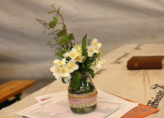 Ein Glasvase mit weißen Blumen steht auf einem Tisch mit Papieren. Dahinter sind ein braunes Buch und ein weißes Tuch zu sehen.