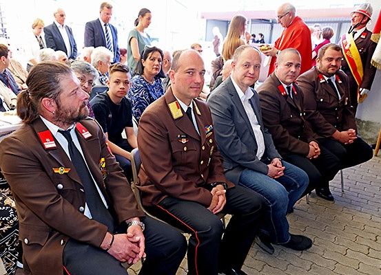 Eine Gruppe formell gekleideter Männer sitzt auf Stühlen bei einer Veranstaltung, mit einem Publikum und Fahnen im Hintergrund.