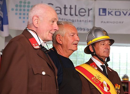 Drei Männer in Uniformen stehen vor einem Banner mit 'Sattle' und 'FARMSCHULE KOVA' darauf. Der Mann auf der linken Seite trägt eine Militäruniform, der Mann in der Mitte trägt ein schwarzes Shirt und der Mann auf der rechten Seite trägt einen Feuerwehrhelm.