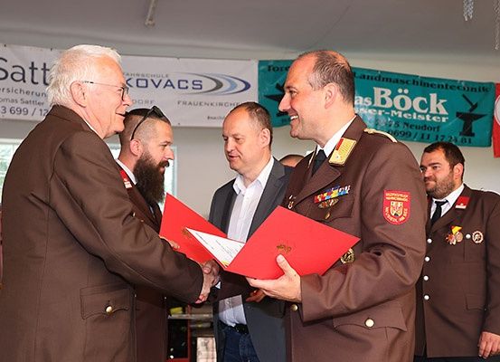 Eine Gruppe von Männern in Militäruniformen und Geschäftskleidung versammelt sich in einem Raum, möglicherweise für eine formelle Veranstaltung. Zwei Männer in der Mitte schütteln sich die Hände und halten eine rote Mappe. Hinter ihnen stehen zwei weitere Männer in Militäruniformen. An der Wand befinden sich Banner mit den Texten 'Satt', 'Frauenkirchen' und 'Bock'.
