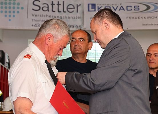 Ein Mann in weißer Uniform wird von einem Mann in einem grauen Anzug geehrt. Dahinter beobachten drei Männer. Die Szene spielt sich vor einem Banner ab.