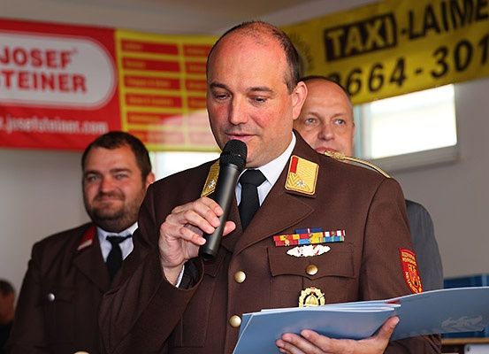 Ein Mann in Uniform spricht in ein Mikrofon und hält Papiere in den Händen. Hinter ihm stehen zwei weitere Männer in Uniform. Ein Banner mit dem Text Josef Steiner ist sichtbar.
