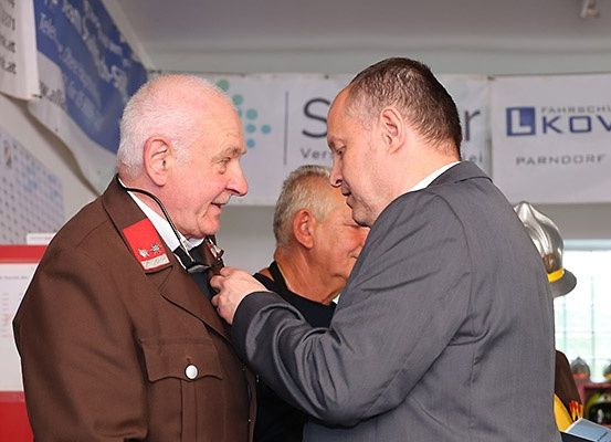 Ein Mann in einer braunen Uniform wird von einem Mann in einem grauen Anzug ausgezeichnet. Hinter ihnen beobachtet ein weiterer Mann. Sie befinden sich in einem Innenbereich mit Bannern.