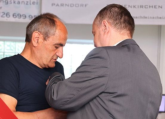 Zwei Männer stehen eng beieinander. Der Mann im schwarzen T-Shirt schaut den Mann im grauen Anzug an. Sie stehen vor einem weißen Banner mit dem Text 'Parndorf'.