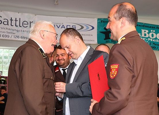 Drei Männer in Uniformen, einer in einem Anzug, führen eine Diskussion. Sie halten eine rote Mappe. Hinter ihnen befinden sich Banner mit den Texten 'Sattler' und 'For'.