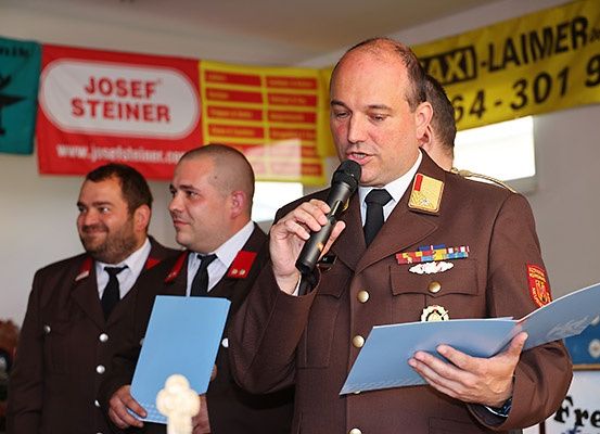 Ein Mann in Uniform spricht in ein Mikrofon bei einer Zeremonie und hält eine blaue Mappe. Zwei Männer in Uniform stehen hinter ihm, ebenfalls mit blauen Ordnern. Ein Banner mit dem Namen Josef Steiner ist sichtbar.