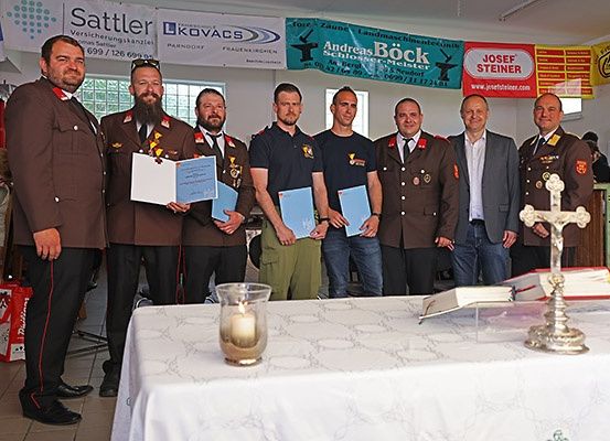 Mehrere Männer in Uniformen stehen vor einem Tisch mit einer Kerze und Büchern. Sie halten Urkunden in den Händen. Ein Mann trägt eine Sonnenbrille auf dem Kopf.