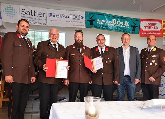 Fünf Männer in Uniform stehen nebeneinander vor einem Tisch und halten Urkunden in den Händen. Dahinter hängen Banner mit den Namen Sattler, L-Kovaks, Andreas Bock und Josef Steiner.