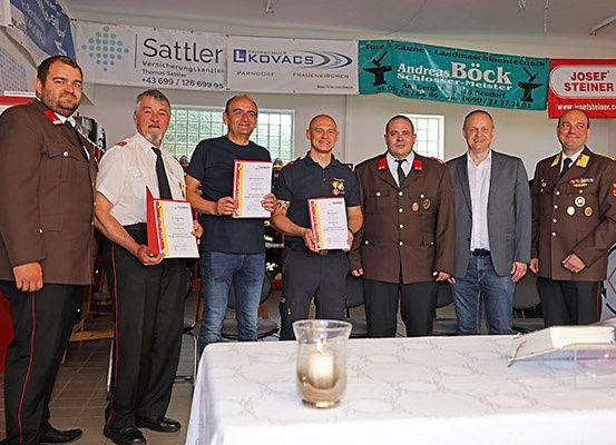 Sechs Männer in Uniformen stehen in einem Raum mit einem Tisch vor ihnen. Sie halten Urkunden in den Händen und lächeln für die Kamera. Hinter ihnen hängt ein Banner an der Wand.