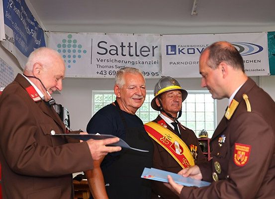 Drei Männer stehen in einem Raum, zwei in Uniformen und einer in einem Feuerwehrhelm. Sie schauen auf Papiere. Hinter ihnen hängen Banner, eines davon trägt die Aufschrift 'Sattler'.