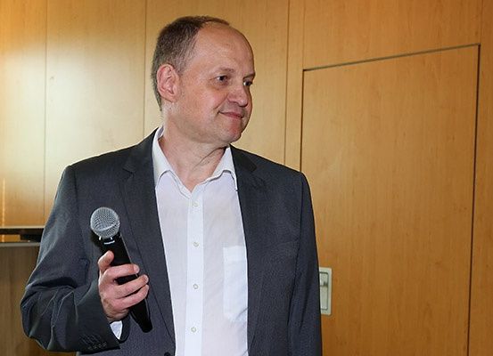 Ein Mann in einem dunklen Anzug hält ein Mikrofon und lächelt in die Kamera. Er steht vor einer Holzwand.