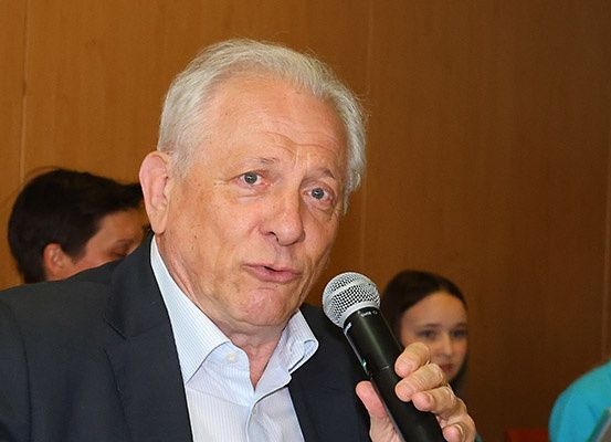 Ein älterer Mann im Anzug spricht in ein Mikrofon und sieht ernst aus, mit verschwommenen Personen im Hintergrund.
