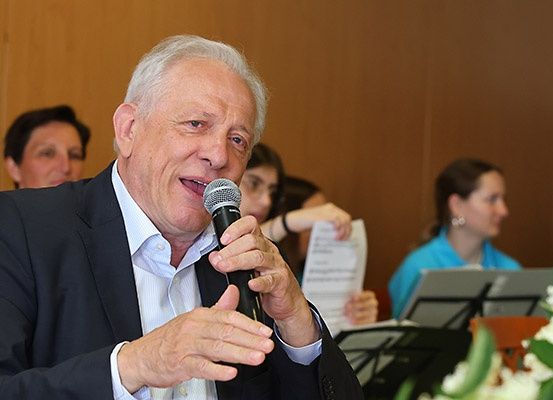 Ein älterer Mann in einem Anzug spricht bei einer Veranstaltung in ein Mikrofon, umgeben von anderen Personen.