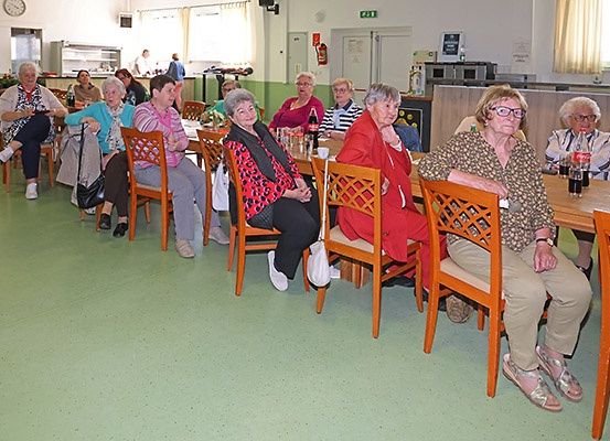 Eine Gruppe älterer Frauen sitzt an Tischen in einem Speisesaal, einige tragen Brillen und Schuhe, mit einem Tisch voller Getränke und einer Person, die in der Nähe der Theke steht.