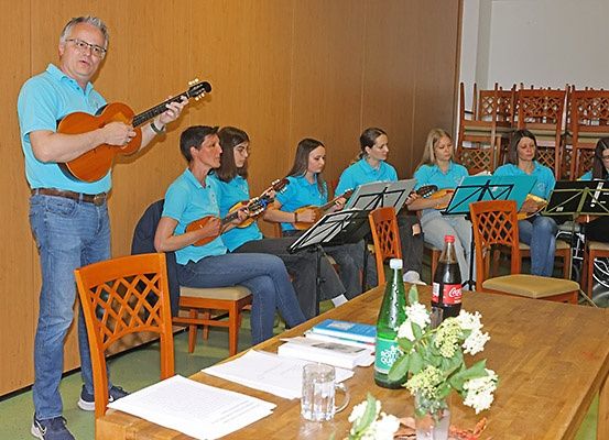 Ein Mann spielt Gitarre, während mehrere Frauen mit Gitarren und Notenblättern vor einem Tisch mit Blumen und Getränken sitzen.