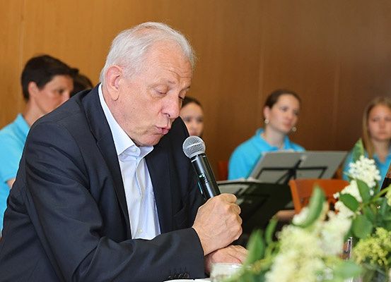 Ein älterer Mann in einem dunklen Anzug spricht in ein Mikrofon und sitzt an einem Tisch mit Blumen. Hinter ihm sitzen mehrere Frauen in blauen Shirts, eine hält ein Buch.