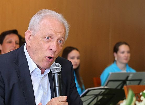 Ein älterer Mann in einem Anzug spricht in ein Mikrofon, während zwei Frauen ihn im Hintergrund betrachten.