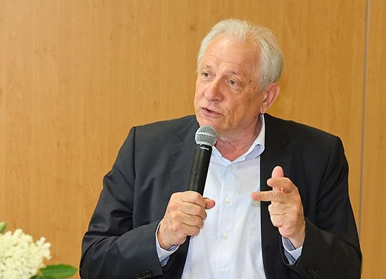 Ein älterer Mann spricht in ein Mikrofon und zeigt mit der Hand, in einem Konferenzraum mit einer hölzernen Wand und einer weißen Blumenanordnung im Vordergrund.