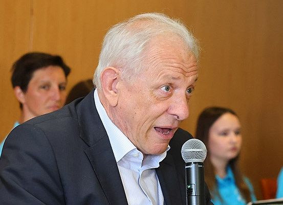 Ein älterer Mann in einem schwarzen Anzug spricht in ein Mikrofon, möglicherweise auf einer Konferenz. Hinter ihm sieht eine Frau in einem blauen Shirt zu ihm.
