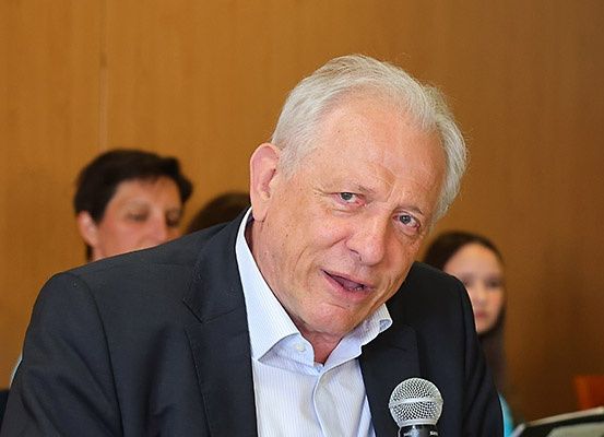 Ein älterer Mann in einem schwarzen Anzug und einem hellblauen Hemd spricht in ein Mikrofon. Eine Frau im Hintergrund ist verschwommen.