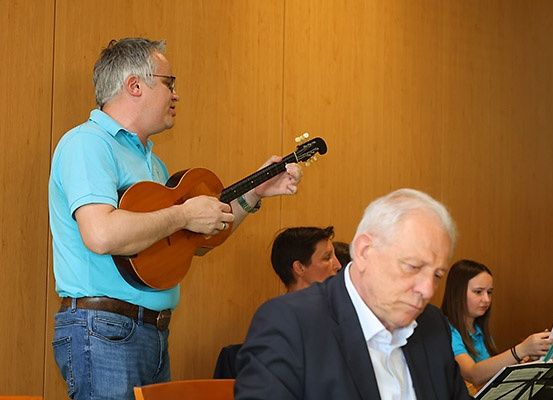 Ein Mann spielt Gitarre und steht vor einer Gruppe. Ein anderer Mann und eine Frau sitzen und schauen Noten an. Der Hintergrund ist eine Holzwand.